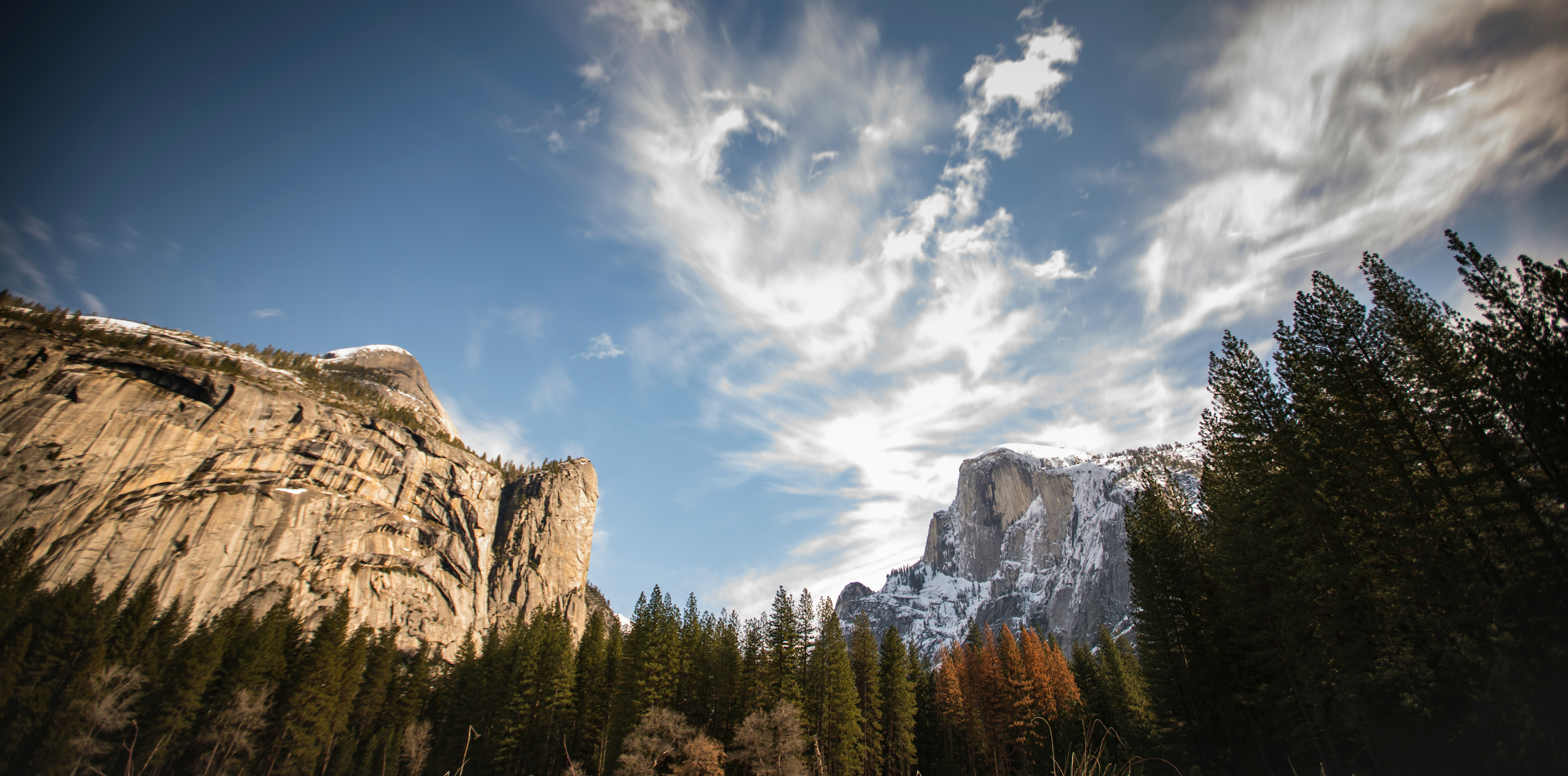 green pine trees near brown rocky mountain under blue and white sunny cloudy sky during daytime